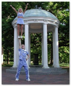 Dr. Landau in a UNC Chapel Hill cheerleading uniform. One of his hands his raised above his head and another UNC cheerleader is standing on his hand with her arms raised. The Old Well rotunda is in the background.