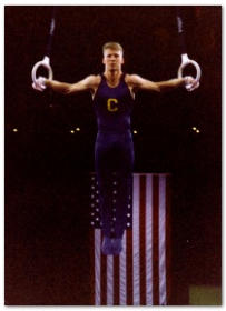 Dr. Joshua Landau competing at the rings in a gymnastics competition with an American flag hanging in the background.