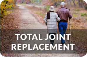 An elderly couple walks arm in arm down an outdoor path in the fall. The man is using a cane to assist him. Title reads: Total Joint Replacement