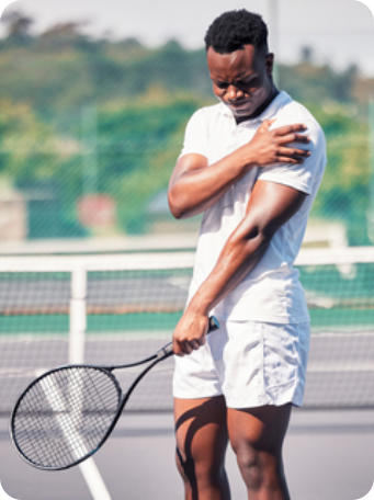 A young man stands on a tennis court holding a racquet in his left hand. His right hand is gripping his left shoulder in pain.