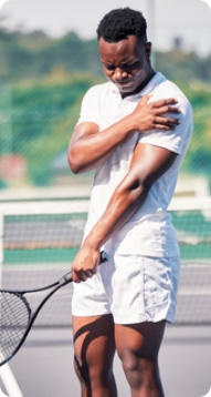 A young man stands on a tennis court holding a racquet in his left hand. His right hand is gripping his left shoulder in pain.