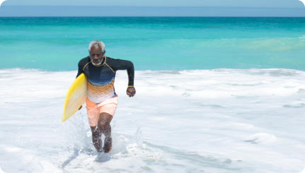 A senior man runs out of the waves on to the beach with a surfboard tucked under his arm.