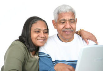A senior couple is sitting together. The woman has her arm around the man's shoulder. They are both looking at a laptop screen and smiling.