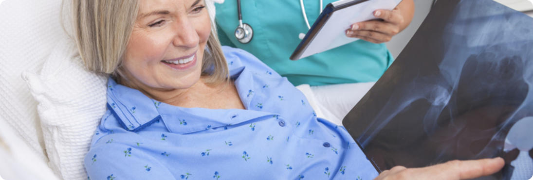 A patient is sitting in a hospital bed and smiling. Her doctor is showing her an x-ray of her successful hip replacement.